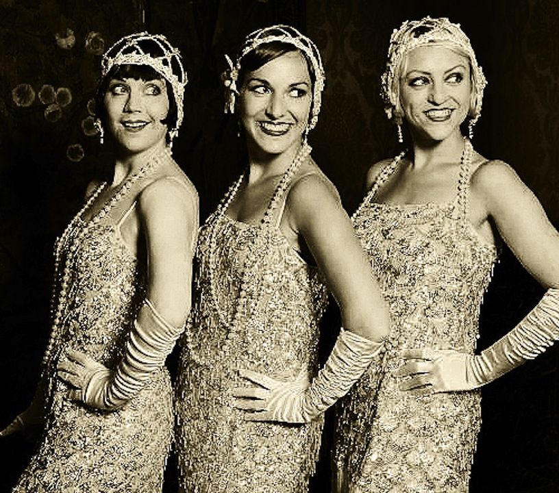 Three women in vintage flapper dresses pose together with smiles.